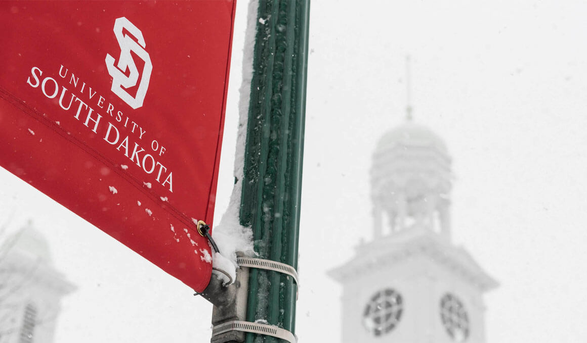 A red USD banner is attached to a light pole during snowfall, with the Old Main tower visible in the blurred background.