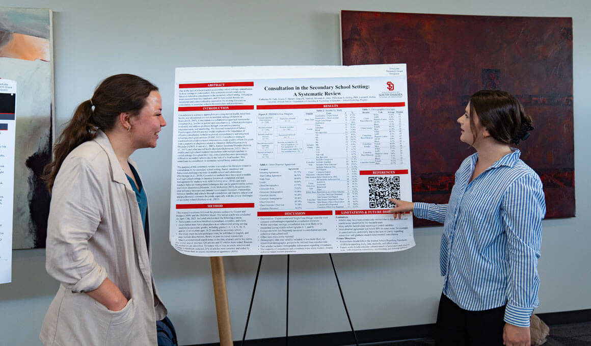A USD student presents and points her poster to a viewer while in a conference area. The poster shows research on a consultation in secondary schools.
