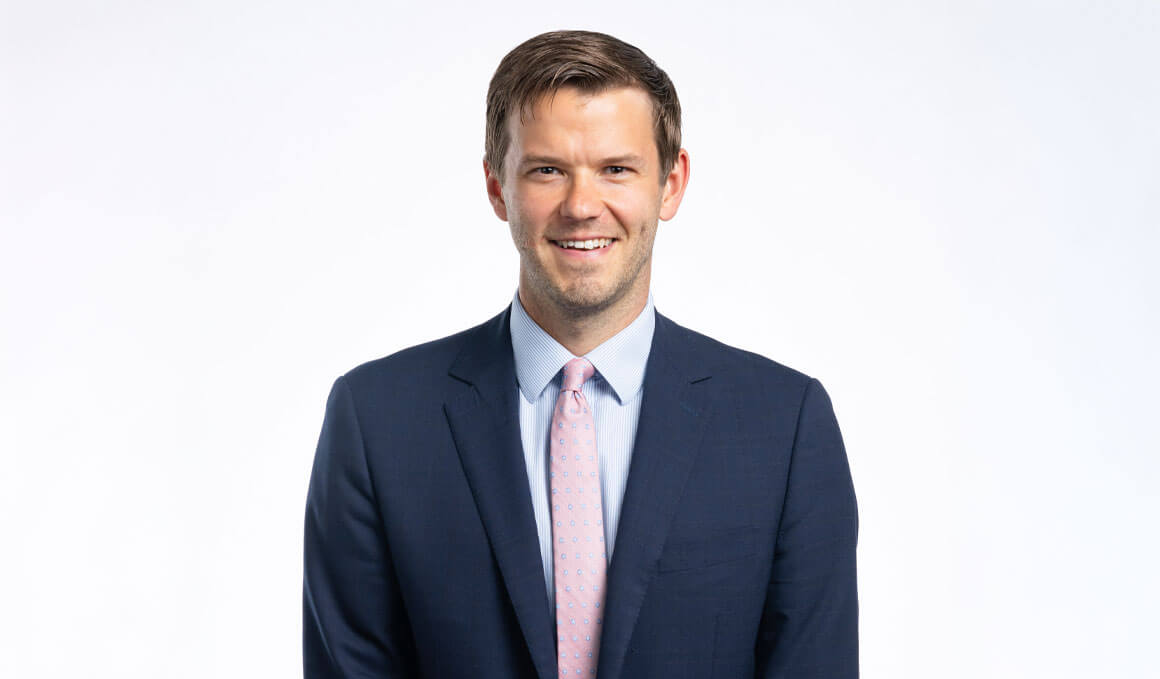 Chris Schmidt, J.D., assistant professor at the University of South Dakota’s Knudson School of Law, smiles at the camera for his professional photo. He is wearing a blue suit with a salmon colored tie and light blue undershirt.