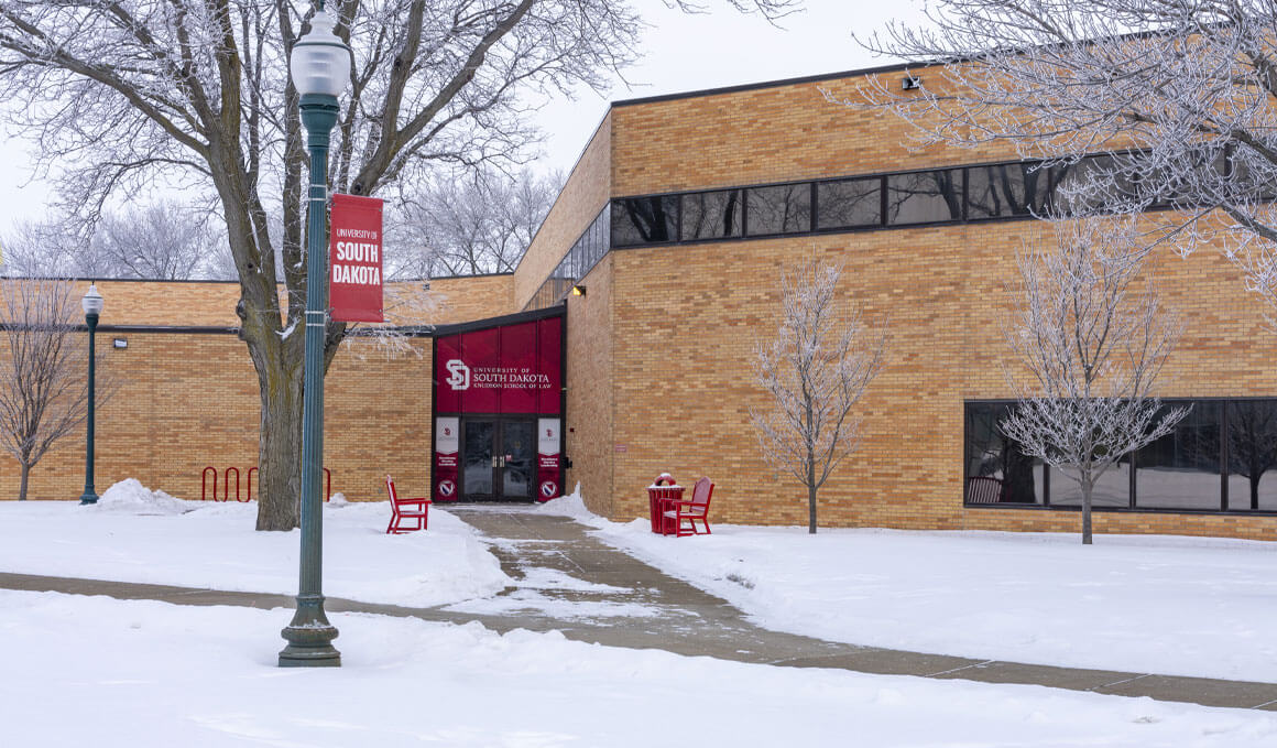 The exterior of the USD Knudson School of Law building on a winter day, with snow-covered ground, bare trees, red benches and a campus banner near the entrance.
