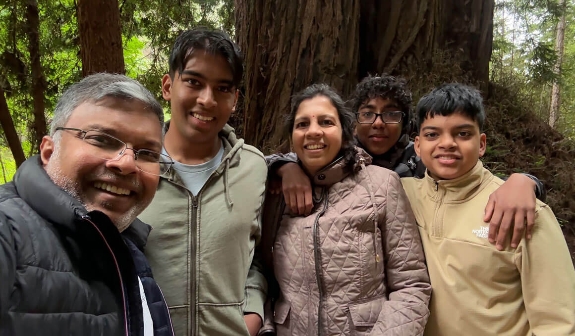 Dr. Thav Thambi-Pillai, far left, stands with his wife, Abi, an IT programmer, and their three boys. They are standing in front of a scenic landscape with trees. 
