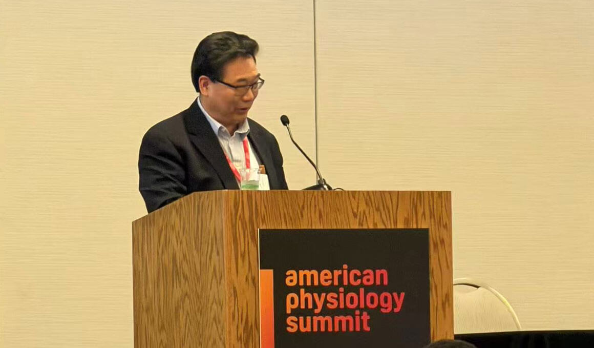 Xuejun Wang stands at a brown, wooden podium, that says "American Physiology Summit." He is speaking into a microphone and wearing a suit.