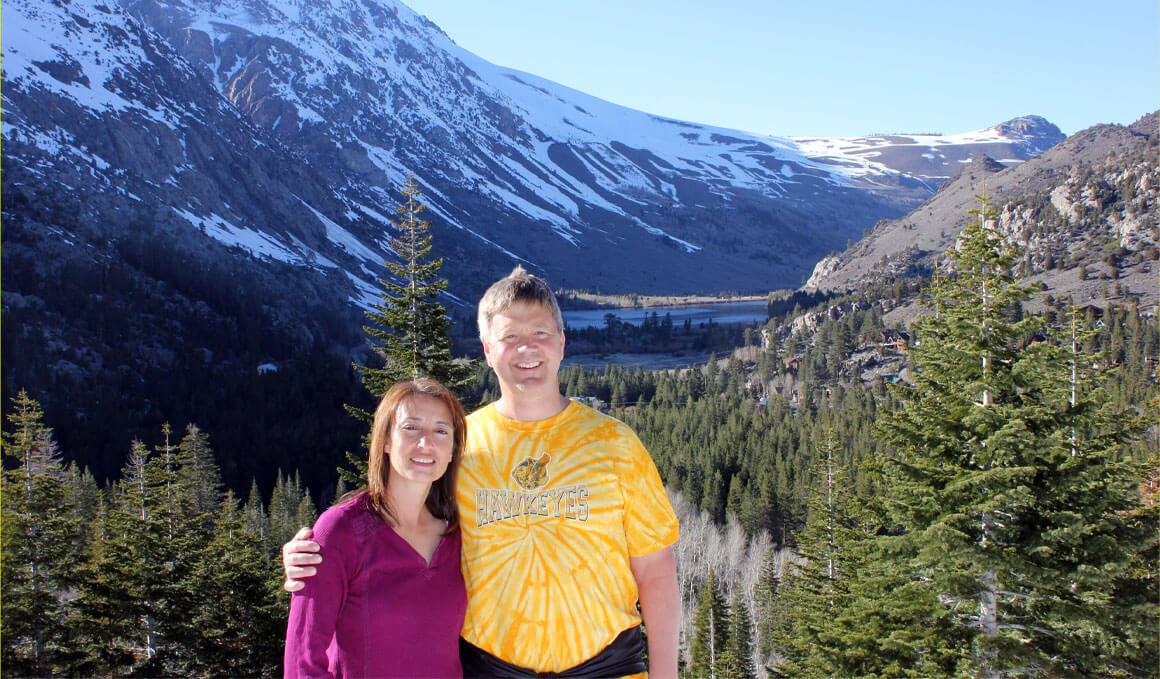 Dr. Lee stands with his wife in front of a beautiful landscape that contains snow-topped mountains and evergreen trees.