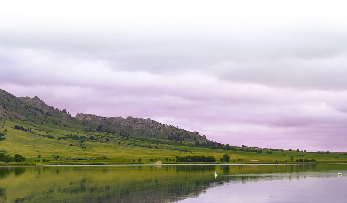 A beautiful scenery of a lake with two birds in it. The lake is surrounded by green grass, trees, and a couple of mountainous hills. The sky behind it is a mix of purple, pink and light bluish-gray.