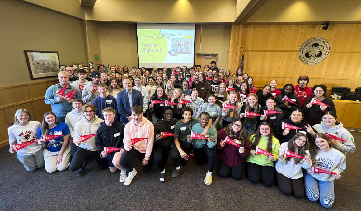 Various high school students from the Sioux Falls School District sit in a conference room, holding USD branded flags. There is a screen in the background that reads "Teacher Flight Path" and there is the SFSD logo on a wall.