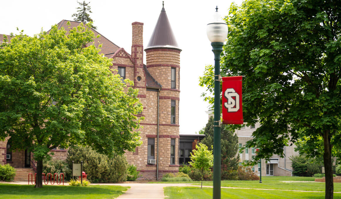 A historic stone building sits on the USD campus, surrounded by trees and grass. There is also a red campus banner on a lamppost. 