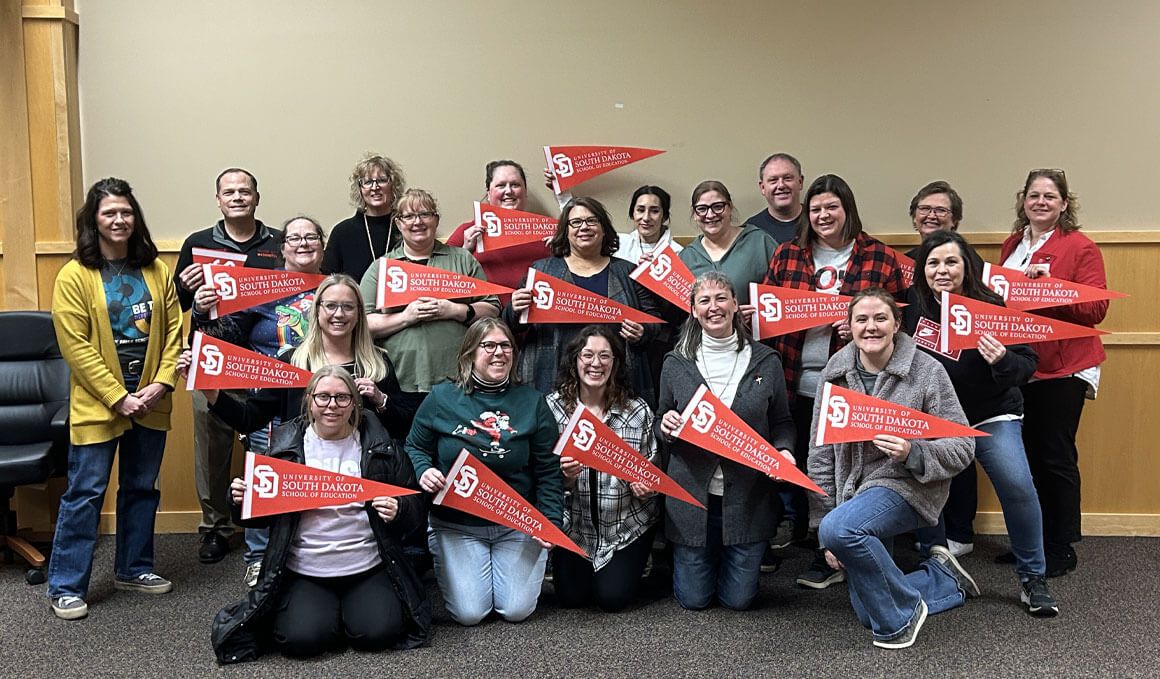 A group of 23 students, who graduated, from the USD and Sioux Falls Professional Development Center, stand in front of a tan wall, posing for a picture. They are holding red flags that are branded with the University of South Dakota logo.