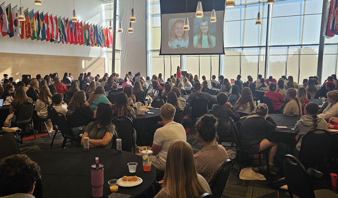 Paige Wright stands on a stage in the ballroom of the MUC, giving a presentation to a big audience, who are seated at round tables.