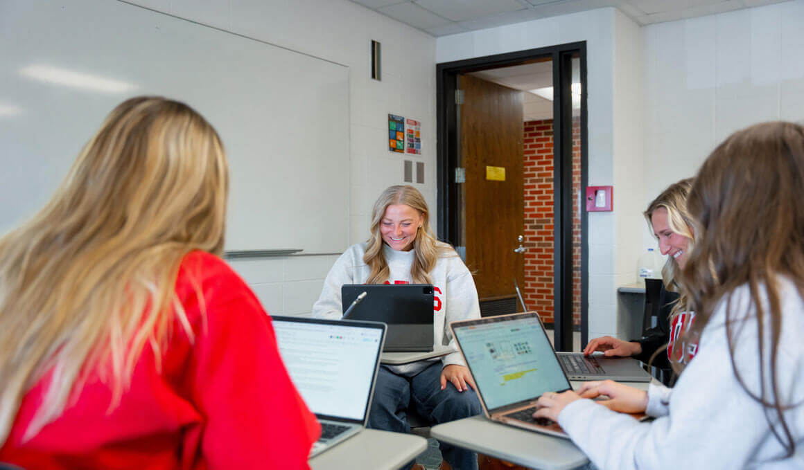 USD students sit in a classroom, while smiling and looking at their laptops. They are sitting in a group-like seating arrangement. A whiteboard and an open door to the classroom are visible in the background.
