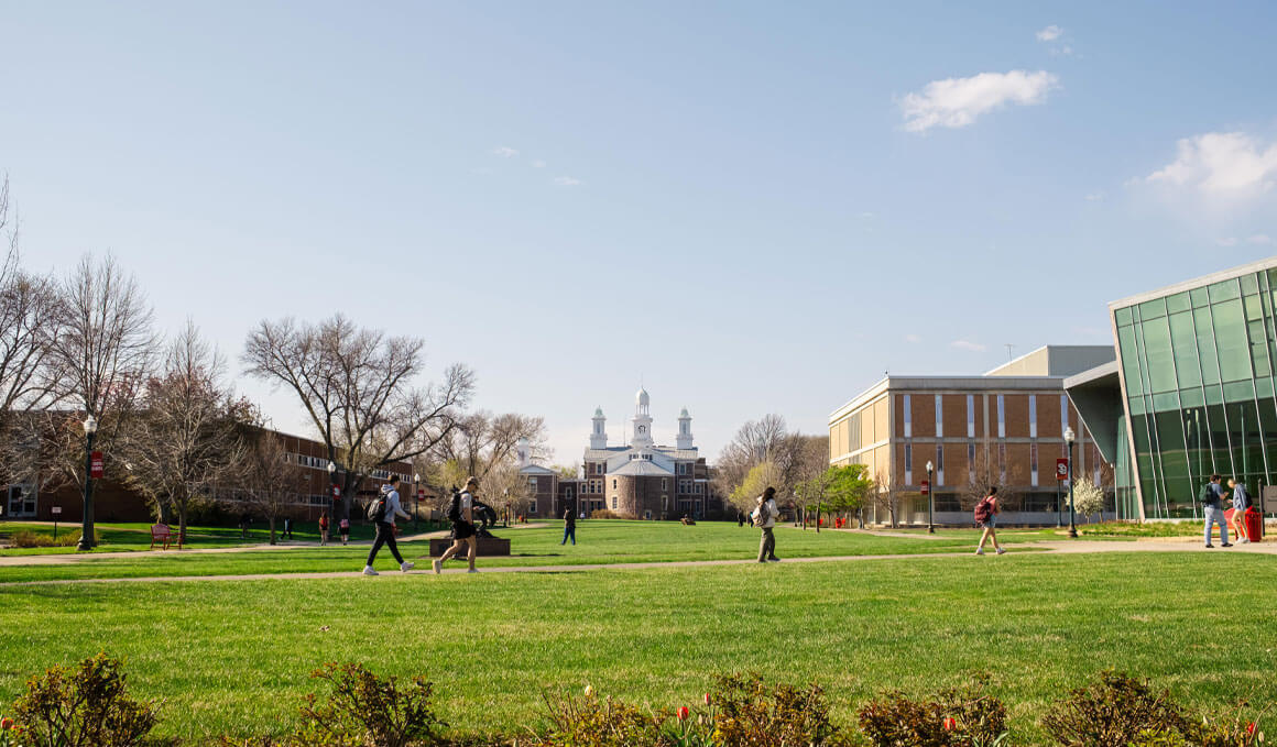 A photo of the main USD campus, focusing on Old Main, with students walking across a grassy quad surrounded by academic buildings on a clear day.