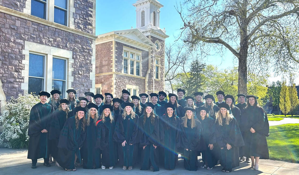 The USD Doctor of Physical Therapy Class of 2026 stands to the side of Old Main for a graduation photo. They are dressed in their graduation regalia and Old Main is slightly visible in the background.
