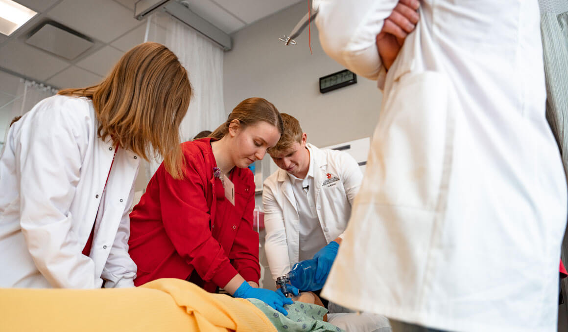 Students in the School of Health Sciences and the Sanford School of Medicine surround a fake patient, using medical instruments to simulate a check-up.