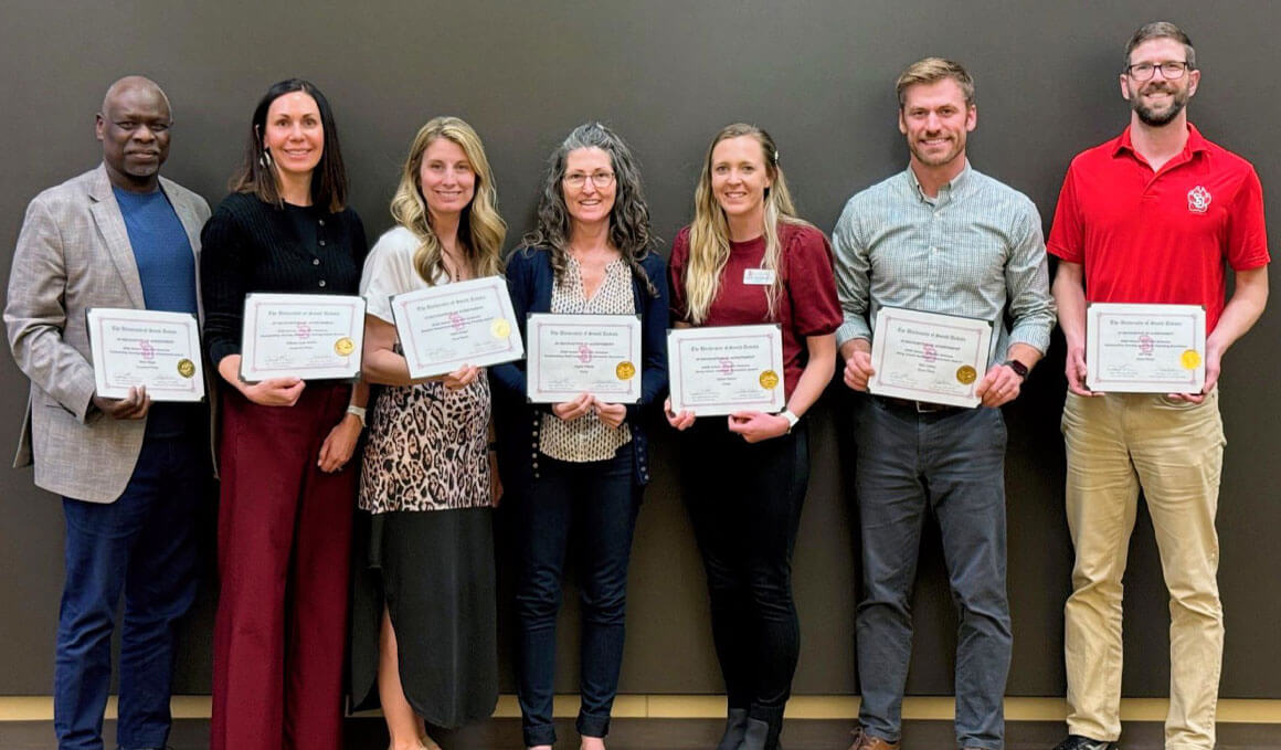 Staff and faculty members from the USD School of Health Sciences stand against a gray wall, holding their certificates. 