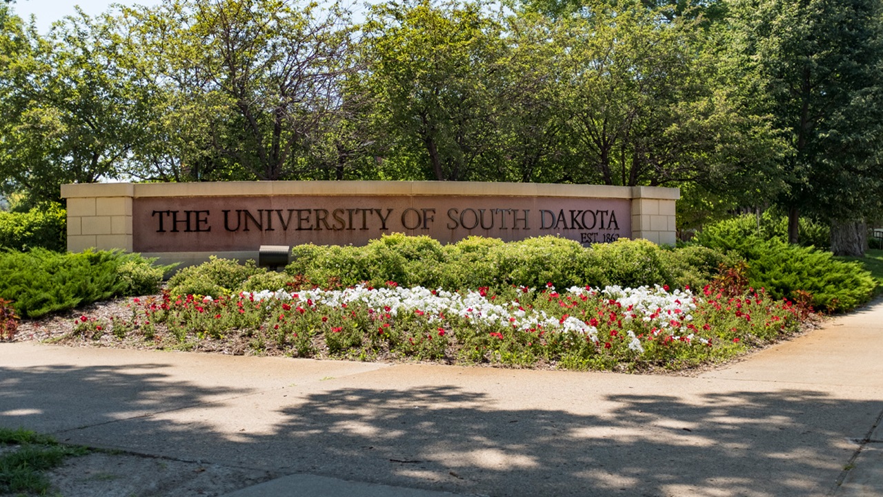 The University of South Dakota stone sign on the corner of campus. There are trees behind the sign and red and white flowers in front of it.