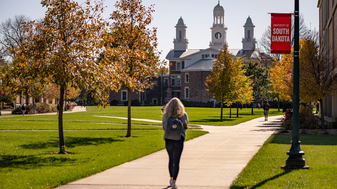 A photo of a student walking on a sidewalk on campus. They are walking towards Old Main.