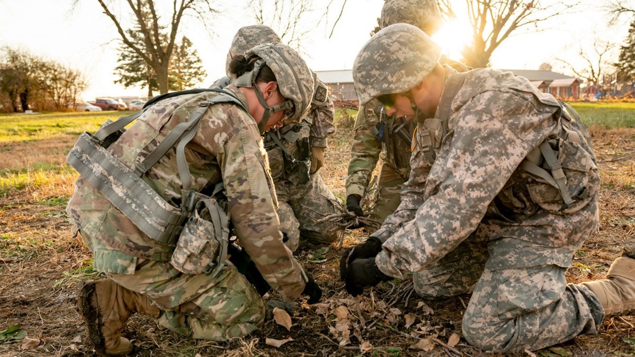 Four people in camouflage military uniforms crouch and huddle around each other on the ground.