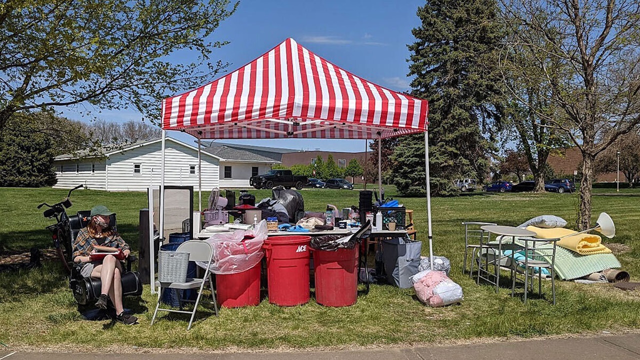A student sits in a chair next to a tent outside. Under the tent, there are many items that students moved out of their dorm rooms to be recycled.