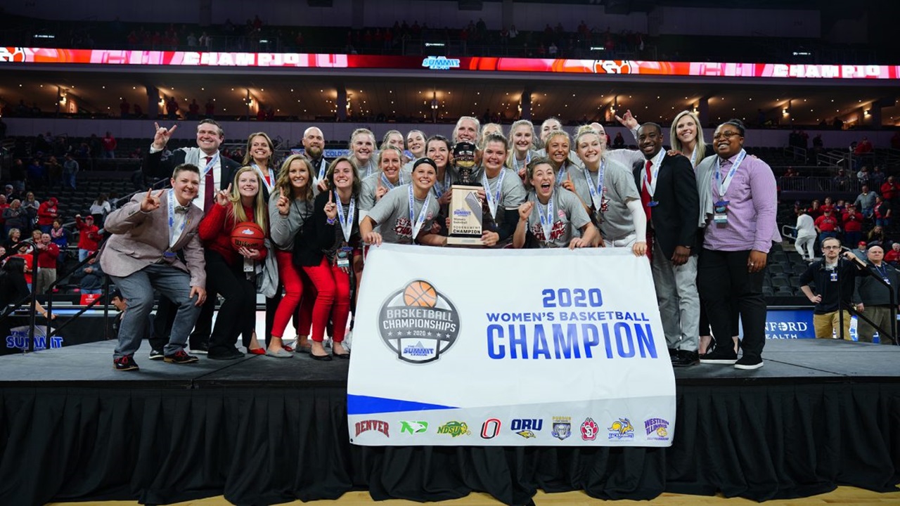 The 2020 USD women's basketball team holds up the 2020 Summit League champions poster and trophy and poses on a stage.