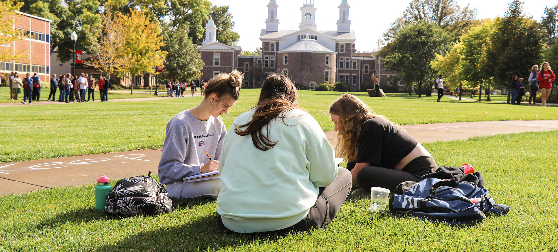 Three students sitting on the MUC lawn doing homework in front of old main.