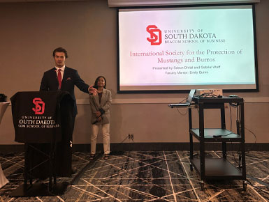Presenter stands at a podium in front of a screen displaying a University of South Dakota Beacom School of Business presentation title, with another person standing nearby.
