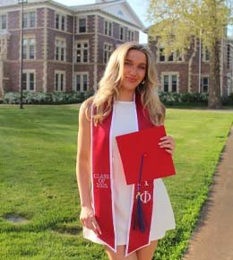 Katie Gellerman poses for a photo in a white dress, with a red graduation sash draped around her, holding her graduation cap.