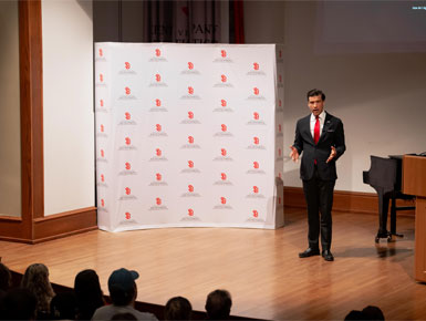 Luke Lindberg stands on stage, speaking to an audience. There is a podium and a piano on his left side, and a branded USD backdrop on his right side. He is wearing a black suit with a bright red tie.