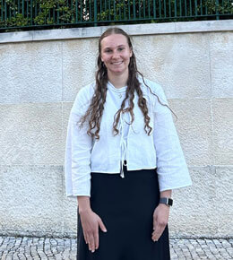 Melanie Brozik stands in front of a brick wall. She is wearing a white shirt and black skirt, smiling at the camera with her hands by her side.