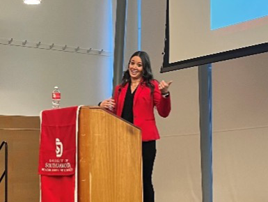 Erica Schilling presents at the Beacom Excellence Dinner. She stands behind a podium and points at a presentation screen behind her.