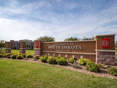 A photo of the brick USD – Sioux Falls sign on the Sioux Falls campus. It is surrounded by blue skies and green grass.