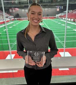 Katie Gellerman stands in the DakotaDome, with the football field behind her, holding the "Outstanding Sorority Senior of the Year Award" from her sorority, Aplha Phi Sorority Psi Chapter.