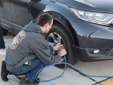A student crouches down next to the front tire on the passenger's side of a blue vehicle. He is holding a hose to an air pump and getting ready to pump the tire with air.