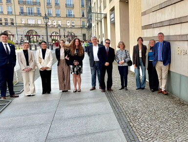 Members of the trade mission trip stand outside a modern office building in a city setting. They are standing on a paved walkway, with historic buildings visible in the background.