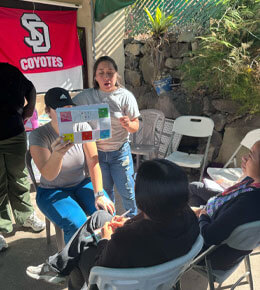An outdoor scene where two USD students are working with Guatemalan community members. One student is seated and holding a printed sheet, while another stands nearby speaking. Several others sit in white folding chairs listening, with a red “Coyotes” banner hanging in the background.