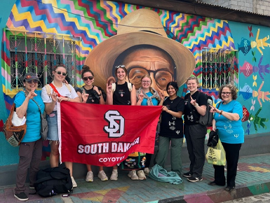 USD students and their chaperones pose with the USD flag in front of a mural in Guatemala.