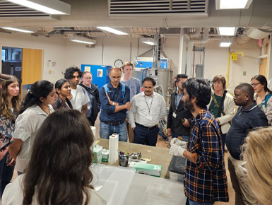 Participants of the Germanium Crystal Growth and Detector Development Lab stand and watch as a faculty member explains the equipment and techniques used in the lab's research.