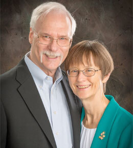 USD alumni Diane Jones Meier, J.D., and Ken Meier, Ph.D., stand together for a professional photo. Ken is wearing a black suit jacket with a blue understhirt and Diane is wearing a teal blazer with a white undershirt. They are smiling at the camera, there is a brown backdrop.