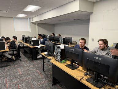 Students sit in front of computer monitors, in a classroom, during the ICPC competition. Some students are smiling and pointing at screens.