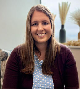 BreAnne Danzi, Ph.D., assistant professor in the USD Department of Psychology, sits on a chair in the CAST Lab, posing for a professional photo. She is wearing a purple cardigan and smiling at the camera.
