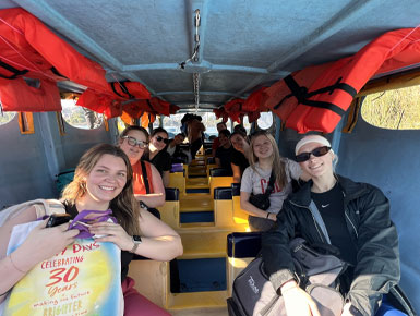 A group of USD students sit inside an open-air boat with rows of yellow benches, with red life jackets anchored above them, smiling and facing the camera during a sunny outing.