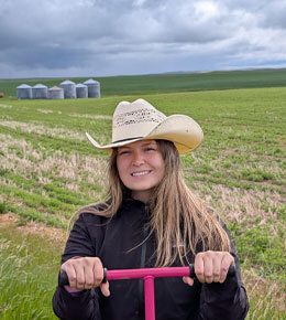 Isabella Rabac stands in a field, wearing a hoodie and a cowgirl hat. Her hands are in front of her, holding two pink handlebars. Behind her is the field and grain bins can be seen in the distant left.