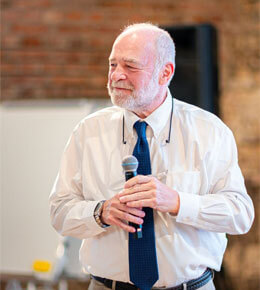 Steve Jarding stands holding a microphone and presenting to an audience. He is wearing a white, button-up shirt and blue tie.