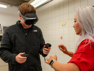 Another USD student tries on a pair of black virtual reality goggles, with help from Professor Young Ae Kim, Ph.D.