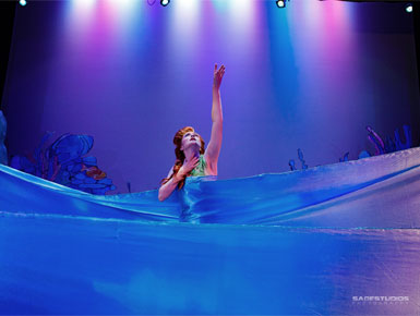 A performer in a blue costume reaches toward colorful stage lights while surrounded by flowing fabric that mimics ocean waves. 