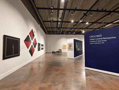 A wide-angle view of the John A. Day Gallery displaying the exhibition "Circle Back: Colleen Cutschall Retrospective" with paintings, a small bed and a navy blue wall partition.