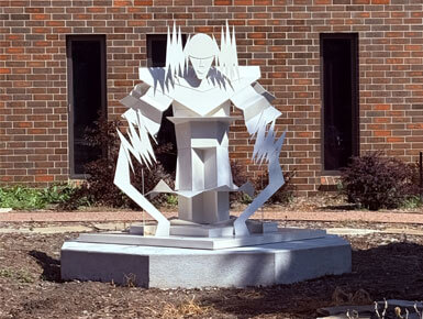 A photo of the Victorious sculpture, which consists of a geometric white metal material standing on a hexagonal stone base in front of a brick building.