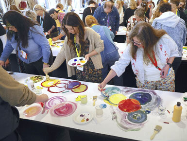 A group of participants are gathered around tables painting with palettes, brushes and colorful circular designs spread across the paper.
