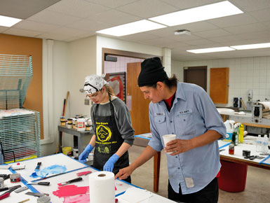 Two artists work together at a studio table covered in blue and red paint during a printmaking session.