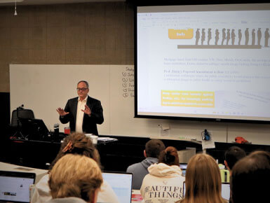 Tom Simmons stands in front of a white board and a smart board, giving a lecture to one of his classes. There are students sitting in front of him, listening intently.