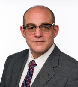 Professional photo of Michael McKey, who is wearing glasses and a grey suit and a striped maroon and blue tie.