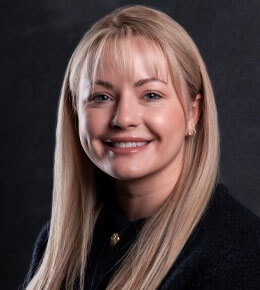 Caitlin Hoeffner smiles at the camera for her professional photo, wearing a dark top against a dark, textured background.